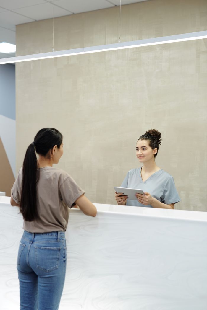 A female receptionist using a tablet to assist a patient at a modern indoor reception desk.
